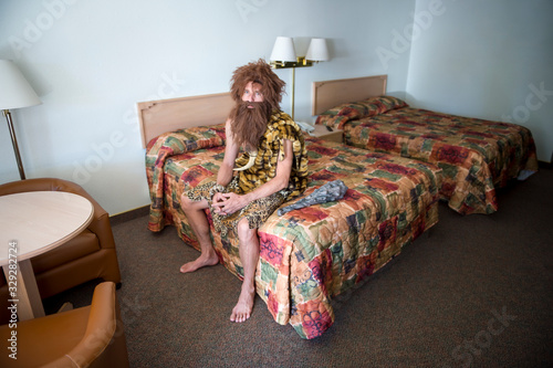 Lonely caveman traveler sitting indoors on the edge of double bed in a dingy motel room
