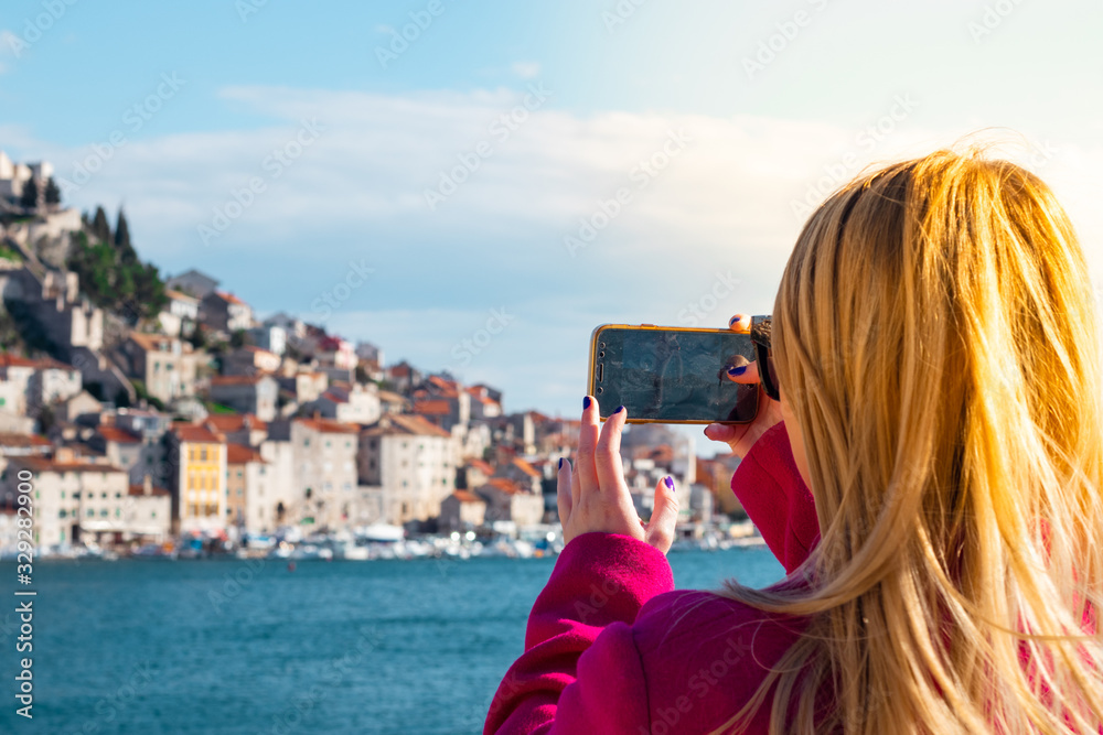 Back view of a blonde woman in a pink coat holding a smartphone taking a photo, town of Sibenik, Croatia in the distance. Blue sea and stone houses