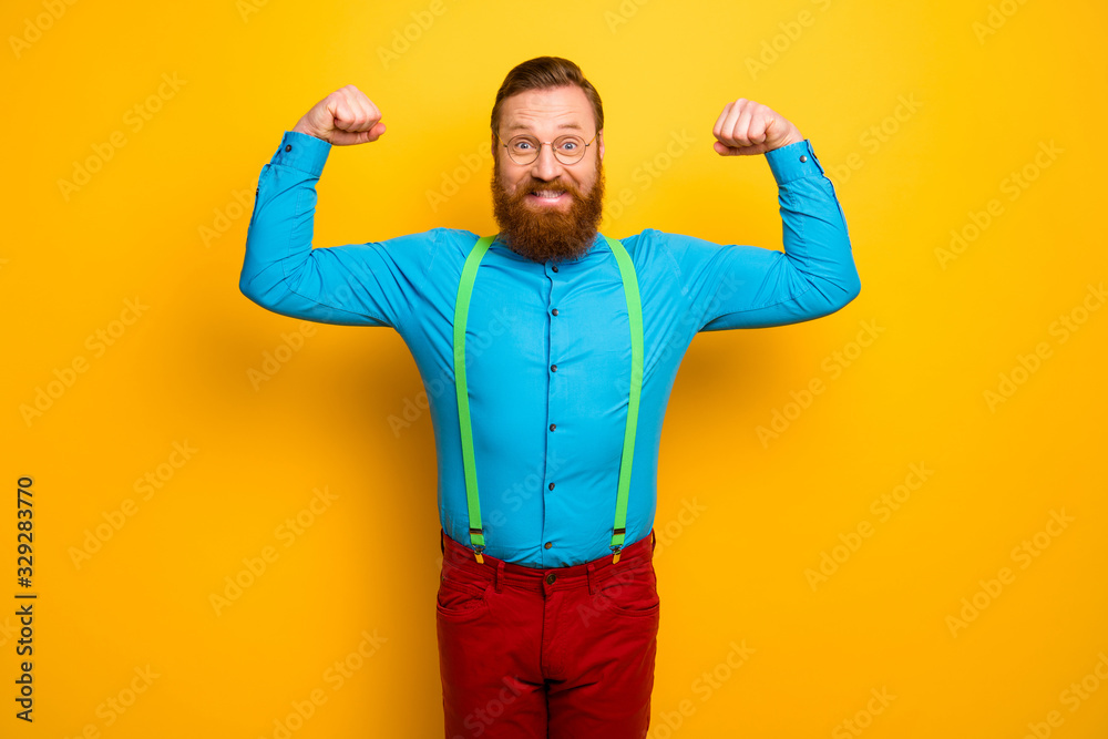 Photo of attractive funny toothy guy raise fists arms presenting perfect shape biceps after training gym wear blue shirt green suspenders red pants isolated yellow color background Photo of attractive funny toothy guy raise fists arms presenting perfect shape biceps after training gym wear blue shirt green suspenders red pants isolated yellow color background