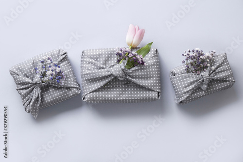 Fototapeta Naklejka Na Ścianę i Meble -  Spring eco-friendly gift wrapped in grey textile with flowers decor on grey. Sustainable gift. Zero waste. Easter holiday. Traditional Japanese Furoshiki style. View from above.