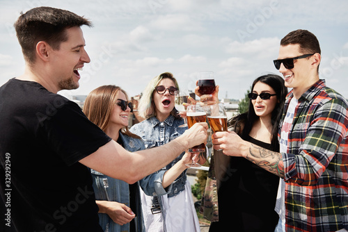 Young happy friends drinking beer before festival at outdoors pub on roof, toasting and laughing, copy space. Friendship and celebration concept