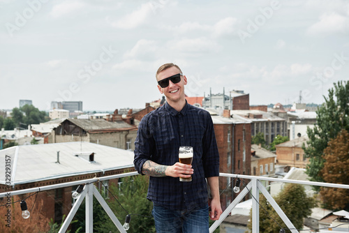 Happy young man in shirt, jeans and eyewear drinking beer at bar or pub on rooftop, copy space