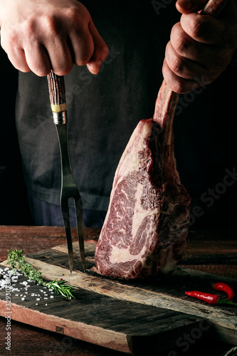 Fényképezés A man holds a Raw Tomahawk steak on a bone of dry aged beef on a dark background