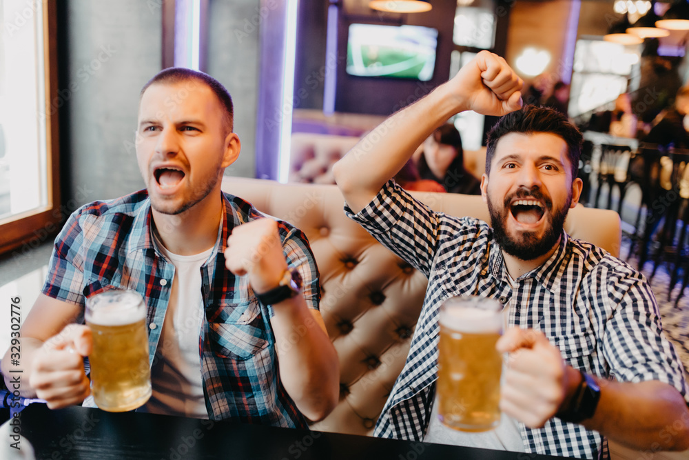 Fans two bearded young man in plaid shirt at bar raised their hands with glasses of beer up and screaming, emotions from exciting game on TV