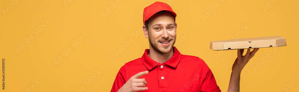 happy delivery man in red uniform pointing with finger at pizza box isolated on yellow, panoramic shot