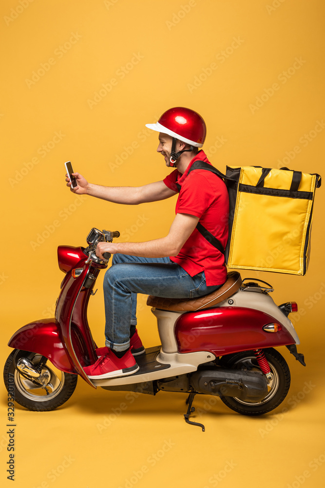 side view of delivery man with backpack on scooter taking selfie on smartphone on yellow background side view of delivery man with backpack on scooter taking selfie on smartphone on yellow background