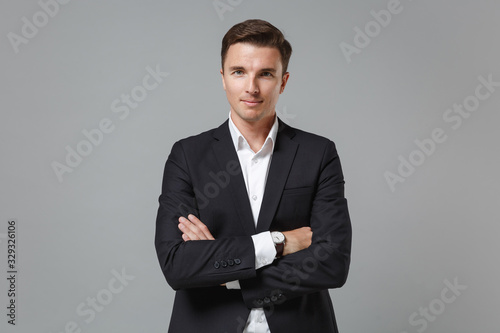 Smiling young business man in classic black suit shirt posing isolated on grey wall background studio portrait. Achievement career wealth business concept. Mock up copy space. Holding hands crossed.