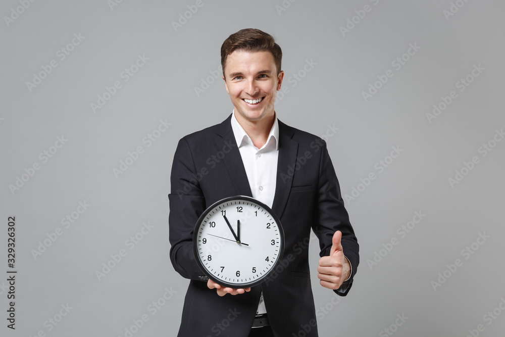 Smiling young business man in classic black suit shirt posing isolated on grey background studio portrait. Achievement career wealth business concept. Mock up copy space. Hold clock, showing thumb up. Smiling young business man in classic black suit shirt posing isolated on grey background studio portrait. Achievement career wealth business concept. Mock up copy space. Hold clock, showing thumb up.