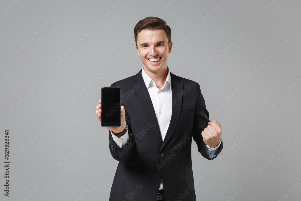 Smiling business man in classic suit shirt isolated on grey background. Achievement career wealth business concept. Mock up copy space. Hold mobile phone with blank empty screen, doing winner gesture. Smiling business man in classic suit shirt isolated on grey background. Achievement career wealth business concept. Mock up copy space. Hold mobile phone with blank empty screen, doing winner gesture.