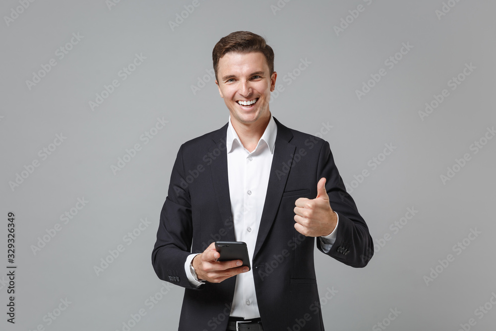 Smiling young business man in classic black suit shirt posing isolated on grey wall background. Achievement career wealth business concept. Mock up copy space. Showing thumb up, hold mobile phone. Smiling young business man in classic black suit shirt posing isolated on grey wall background. Achievement career wealth business concept. Mock up copy space. Showing thumb up, hold mobile phone.