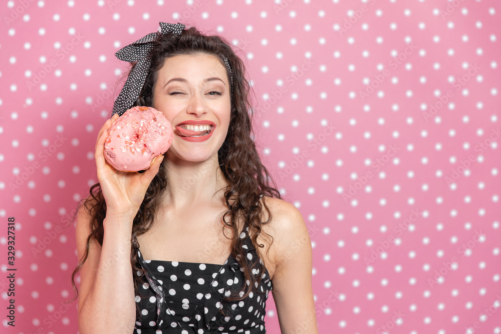 Glamorous young girl is holding yummy doughnut, sticking out her tongue trying to lick out a pink icing and winking at the camera. Glamorous young girl is holding yummy doughnut, sticking out her tongue trying to lick out a pink icing and winking at the camera.