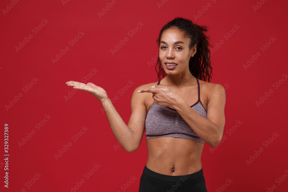 Smiling young african american sports fitness woman in sportswear posing working out isolated on red background in studio. Sport exercises healthy lifestyle concept. Pointing index finger, hand aside. Smiling young african american sports fitness woman in sportswear posing working out isolated on red background in studio. Sport exercises healthy lifestyle concept. Pointing index finger, hand aside.