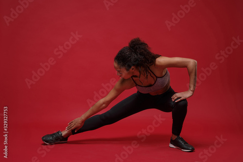 Strong young african american sports fitness woman in sportswear posing working out isolated on red wall background studio portrait. Sport exercises healthy lifestyle concept. Stretching her legs.
