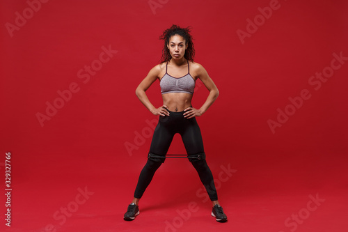 Strong african american sports fitness woman in sportswear working out isolated on red background. Sport exercises healthy lifestyle concept. Stand with arms akimbo, doing exercises with elastic band.