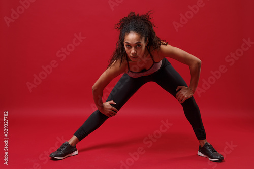 Fototapeta Naklejka Na Ścianę i Meble -  Strong young african american sports fitness woman in sportswear posing working out isolated on red wall background studio portrait. Sport exercises healthy lifestyle concept. Stretching her legs.