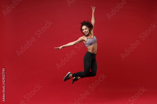 Cheerful young african american fitness woman in sportswear posing working out isolated on red wall background studio portrait. Sport exercises healthy lifestyle concept. Jumping, spreading hands.