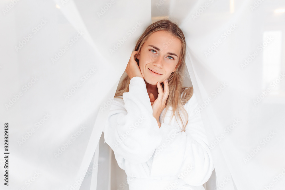 Portrait of pleased young charming smiling woman looking at camera on a white fabric background on sunny morning. ?oncept of positive start day, good mood, Youth and health care, beauty and self-care Portrait of pleased young charming smiling woman looking at camera on a white fabric background on sunny morning. ?oncept of positive start day, good mood, Youth and health care, beauty and self-care