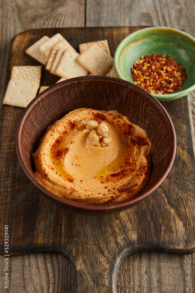 Selective focus of crackers, bowls with paprika and hummus on cutting board on wooden background