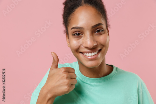Close up of smiling young african american woman girl in green sweatshirt posing isolated on pastel pink background studio portrait. People lifestyle concept. Mock up copy space. Showing thumb up.
