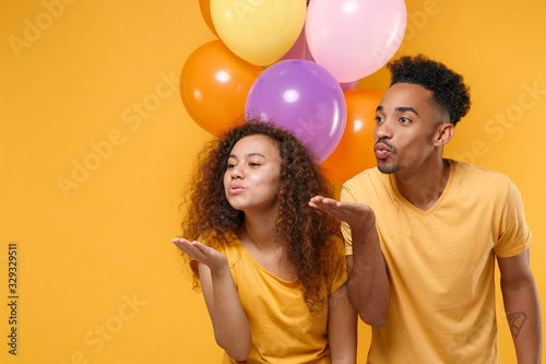Cute friends couple african american guy girl in casual clothes isolated on yellow background. Birthday holiday party lifestyle concept. Celebrate hold colorful air balloons blowing sending air kiss. © ViDi Studio