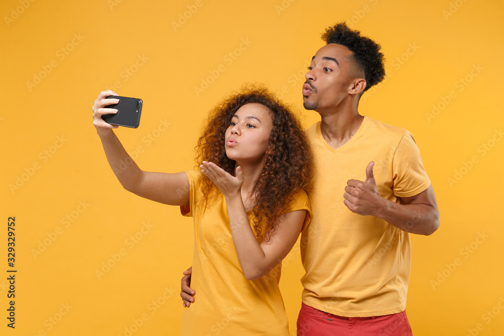Pretty friends couple african american guy girl in casual clothes isolated on yellow background. People lifestyle concept. Mock up copy space. Doing selfie shot on mobile phone blowing send air kiss. Pretty friends couple african american guy girl in casual clothes isolated on yellow background. People lifestyle concept. Mock up copy space. Doing selfie shot on mobile phone blowing send air kiss.