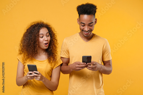 Amazed young friends couple african american guy girl in casual clothes isolated on yellow orange wall background. People lifestyle concept. Mock up copy space. Using mobile phone, typing sms message.