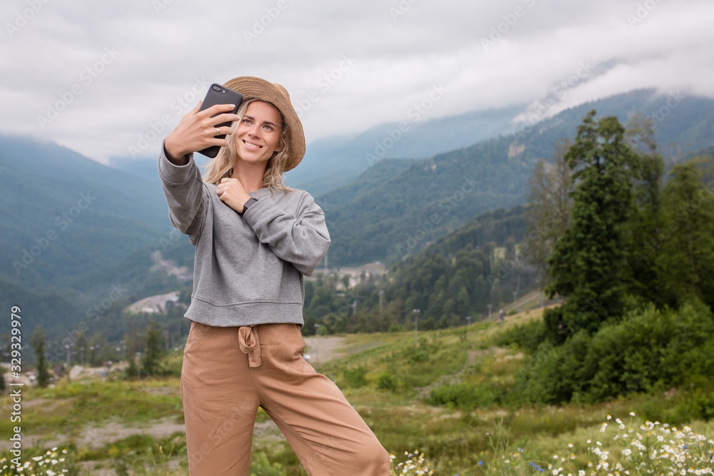 Inspired young caucasian woman makes selfie on smartphone while standing on high peak of mountain. Pretty woman takes photos for social networks while traveling. Vacation conquering peaks concept