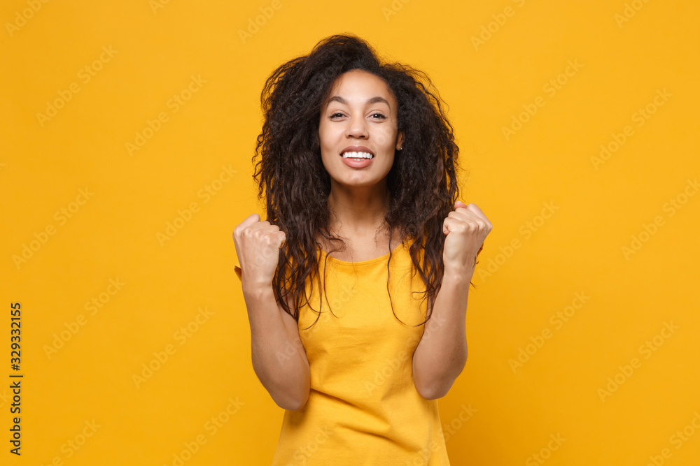 Cheerful young african american woman girl in casual t-shirt posing isolated on yellow orange background studio portrait. People lifestyle concept. Mock up copy space. Clenching fists like winner. Cheerful young african american woman girl in casual t-shirt posing isolated on yellow orange background studio portrait. People lifestyle concept. Mock up copy space. Clenching fists like winner.