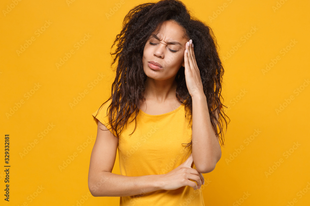 Sick young african american woman in casual t-shirt posing isolated on yellow orange background. People lifestyle concept. Mock up copy space. Put hand on head, keeping eyes closed, having headache.
