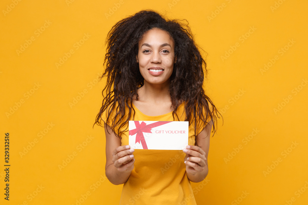 Smiling young african american woman girl in casual t-shirt posing isolated on yellow orange background studio portrait. People emotions lifestyle concept. Mock up copy space. Hold gift certificate. Smiling young african american woman girl in casual t-shirt posing isolated on yellow orange background studio portrait. People emotions lifestyle concept. Mock up copy space. Hold gift certificate.