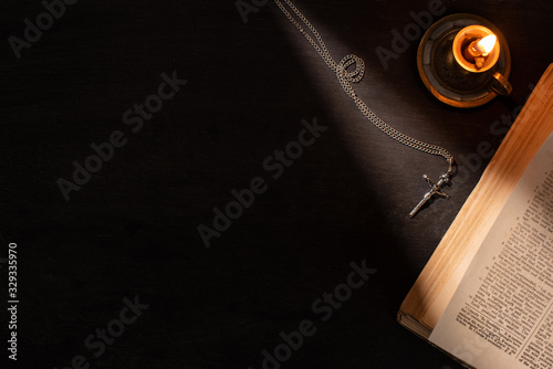 top view of holy bible near burning candle and cross on dark background with sunlight