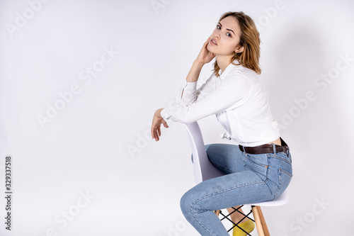 young sexy girl with red hair in a white shirt and jeans sits on a chair on a white background, copy space