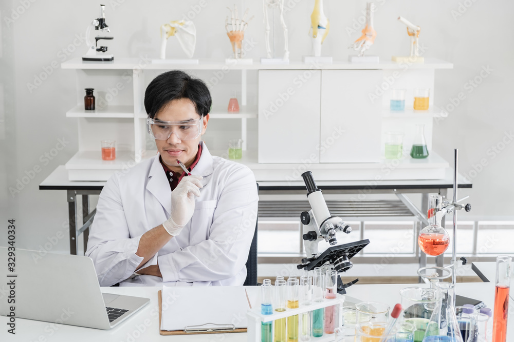 A male scientist with black hair wearing white coat and protective glassware thinking sitting with laptop and microscope in white laboratory room with test tube and solutions.