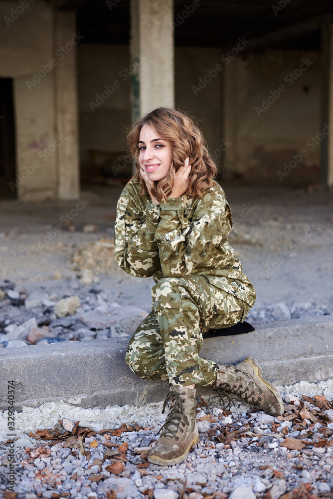 Young curly blond military woman, wearing ukrainian military uniform, sitting on pavement edge, smiling. Full-length portrait of army soldier in front of ruined abandoned building, construction site.