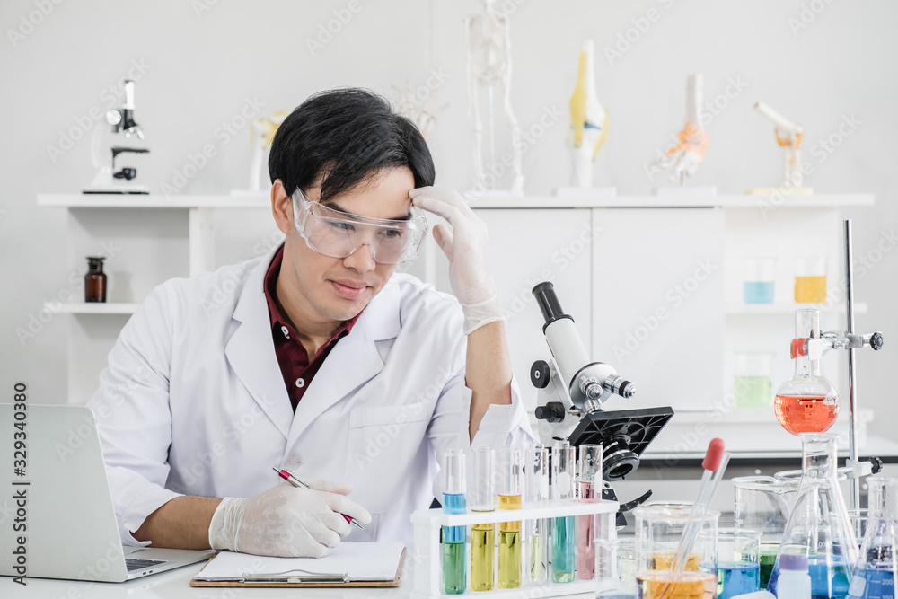 A male scientist with black hair wearing white coat and protective glassware sitting with laptop looking at a microscope in worry in white laboratory room with test tube and solutions. A male scientist with black hair wearing white coat and protective glassware sitting with laptop looking at a microscope in worry in white laboratory room with test tube and solutions.