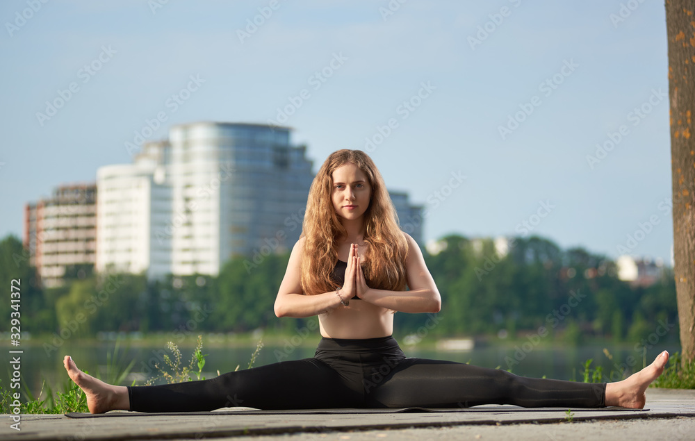 Tender girl with long hair practicing yoga outdoors near the lake. Girl working out in black pants and bra at mat against blurred background of buildings at city. Yoga and healthy lifestyle concept
