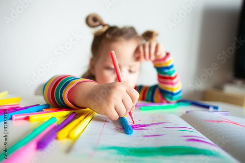 Little girl, a 3 year old girl, with a ponytail hairstyle in a multi-colored colorful striped jacket on a light background at the table draws multicolored markers and smiles