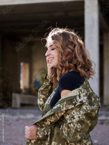 Young curly blond military woman, wearing ukrainian army military uniform and black t-shirt, posing. Three-quarter portrait of female soldier in front of ruined abandoned building,construction site.