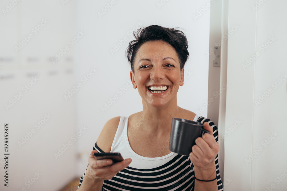 beautiful smiling mid aged woman using mobile phone and holding cup of coffee or tea at her home beautiful smiling mid aged woman using mobile phone and holding cup of coffee or tea at her home