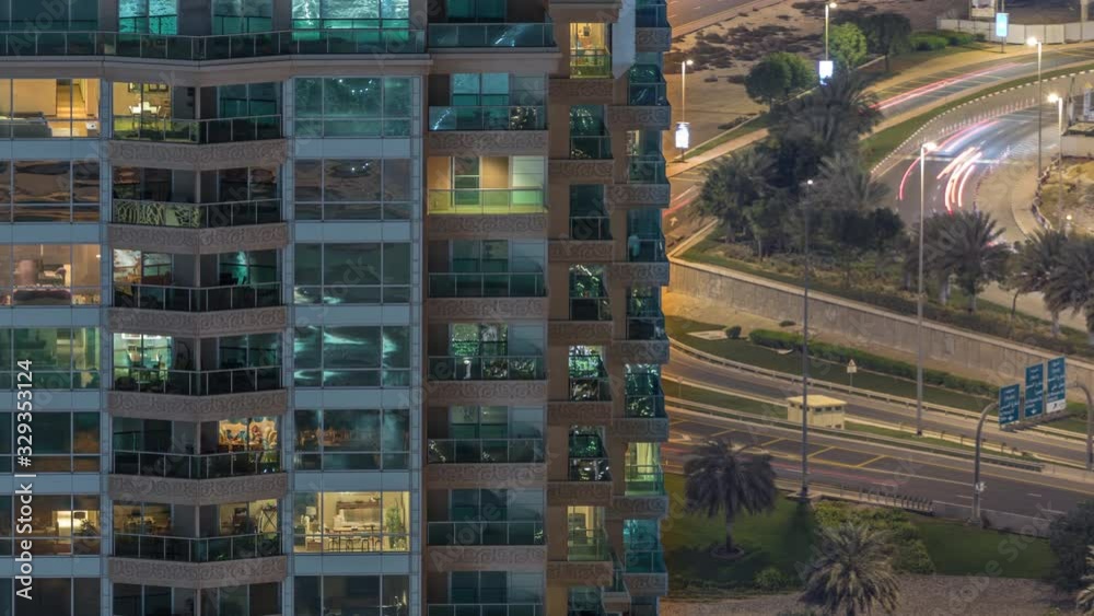 Rows of glowing windows with people in the interior of apartment building at night. Modern skyscraper with glass surface. Traffic on roads on a background. Zoom in Rows of glowing windows with people in the interior of apartment building at night. Modern skyscraper with glass surface. Traffic on roads on a background. Zoom in
