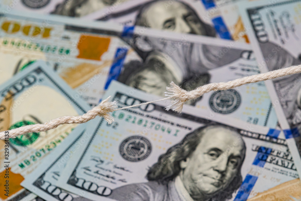 Closeup of frayed breaking rope with 100 dollar bills in background. Concept of fragile economy, stock market crash, and recession Closeup of frayed breaking rope with 100 dollar bills in background. Concept of fragile economy, stock market crash, and recession