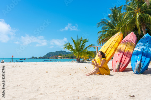 Girl in a yellow dress on a tropical sandy beach works on a laptop near kayaks and drinks fresh mango. Remote work  successful freelance. Works on vacation.