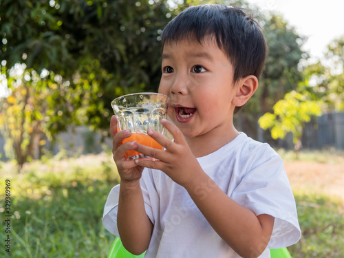Asian little child boy holding glass of orange juice with smiling happy face outdoor nature background at home. Vitamin C nutrition for healthy concept.