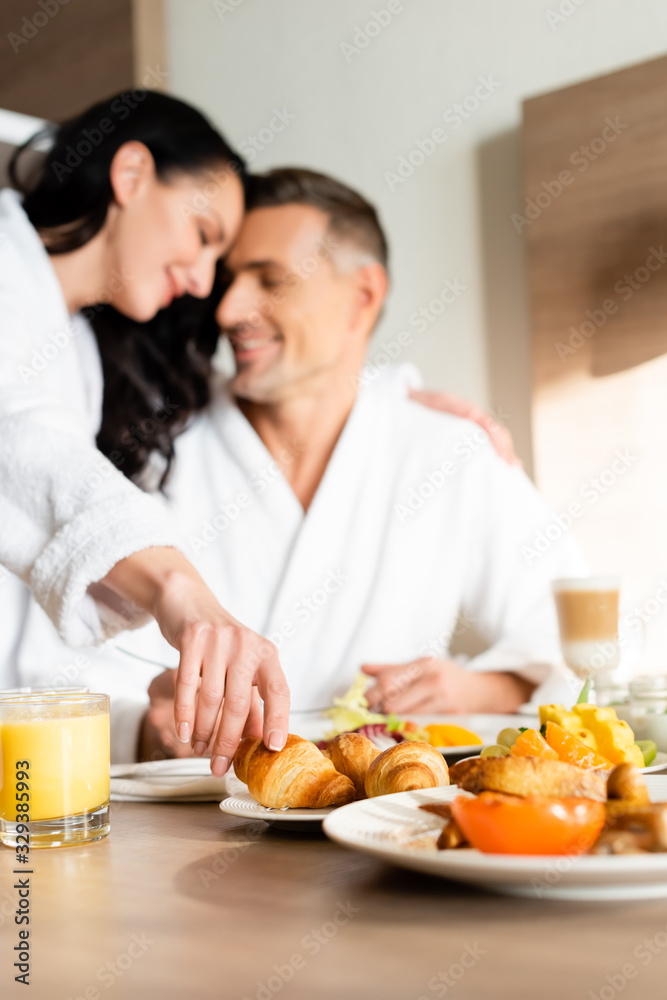 selective focus of smiling girlfriend in bathrobe hugging boyfriend and taking croissant in hotel