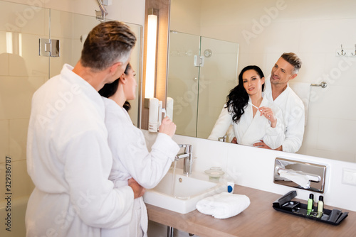 boyfriend hugging smiling girlfriend in bathrobe with toothbrush in hotel