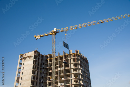 Tower crane lifts the block for building under construction at background blue sky at sunny day. Precast concrete slab hanging from crane hook above building skeleton at construction site