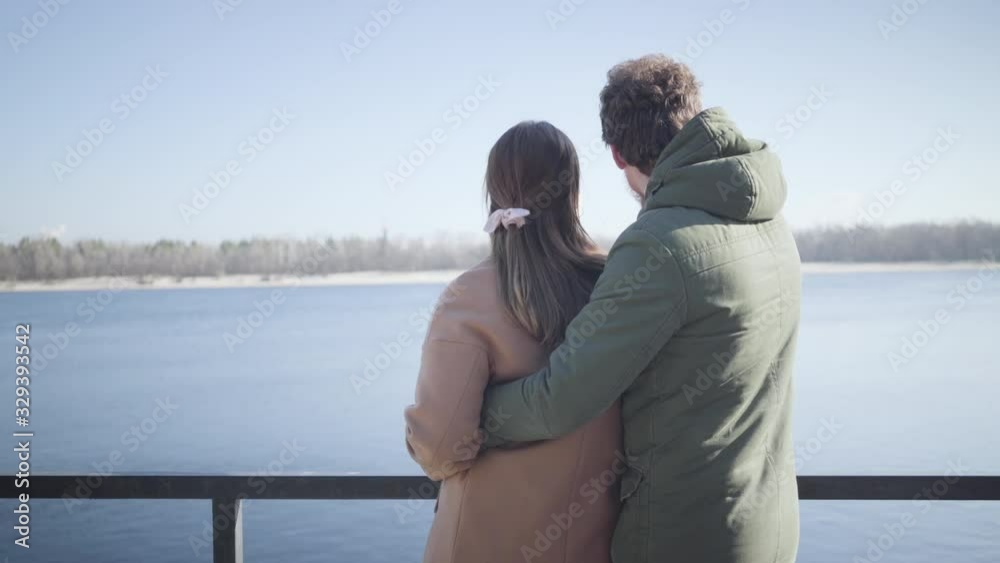 Back view of happy young couple chatting on river bank on sunny autumn day. Caucasian man hugging girlfriend and pointing at something away. Joy, love, romance, lifestyle, unity. Back view of happy young couple chatting on river bank on sunny autumn day. Caucasian man hugging girlfriend and pointing at something away. Joy, love, romance, lifestyle, unity.