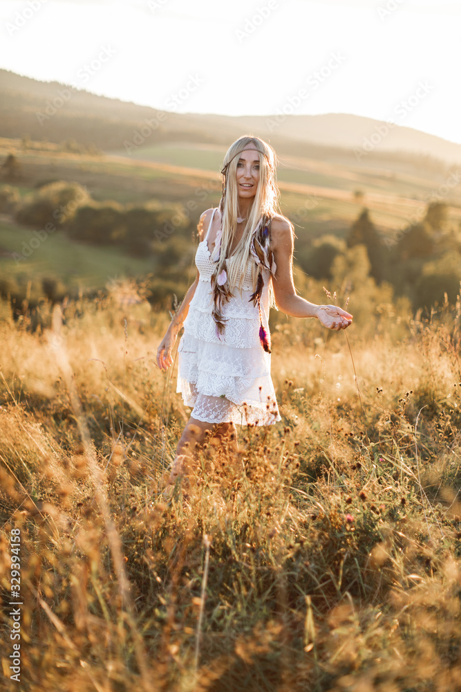 Portrait of happy smiling woman with long blonde hair is wearing white boho hippie dress in field. Feathers in hair, boho style Portrait of happy smiling woman with long blonde hair is wearing white boho hippie dress in field. Feathers in hair, boho style