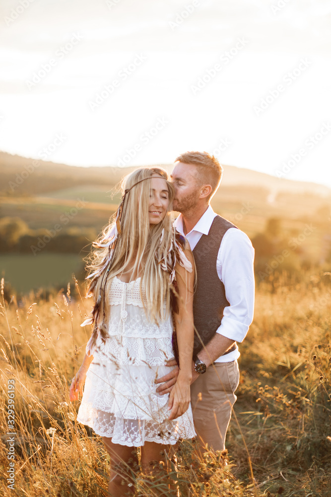 boho couple holding hands and embracing together, walking in countryside field. Pretty woman in white dress and boho feather accessories in hair, handsome man in casual suit