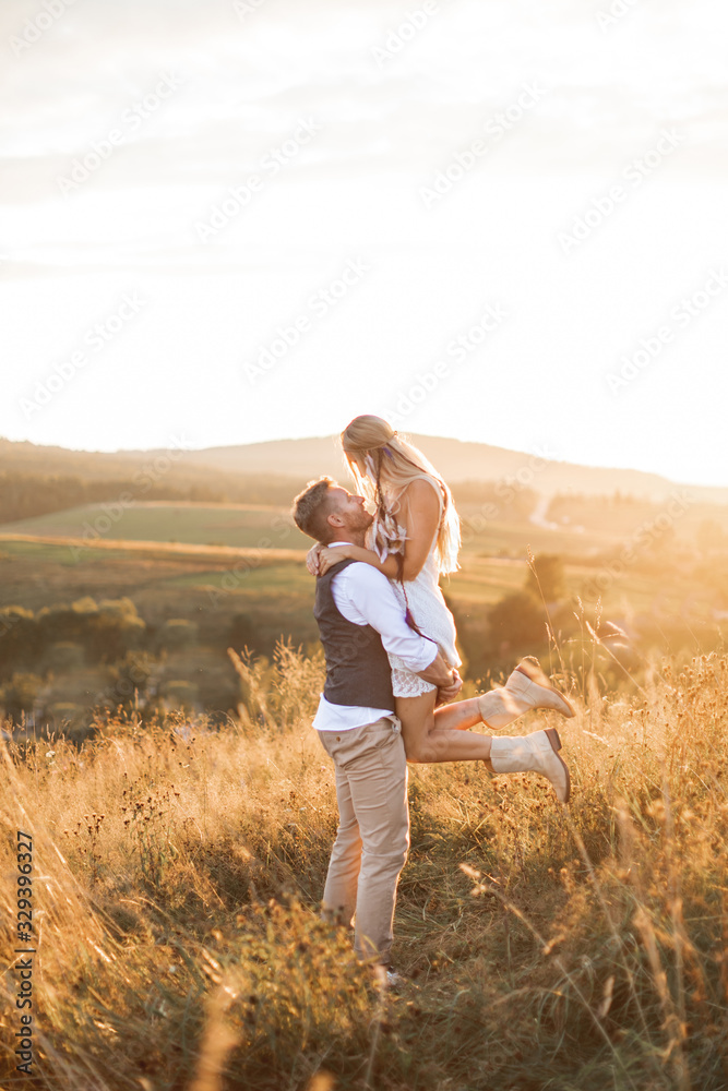 Shot of young boho hippie woman being carried by her handsome boyfriend in summer field. Couple having fun on their summer evening outdoors